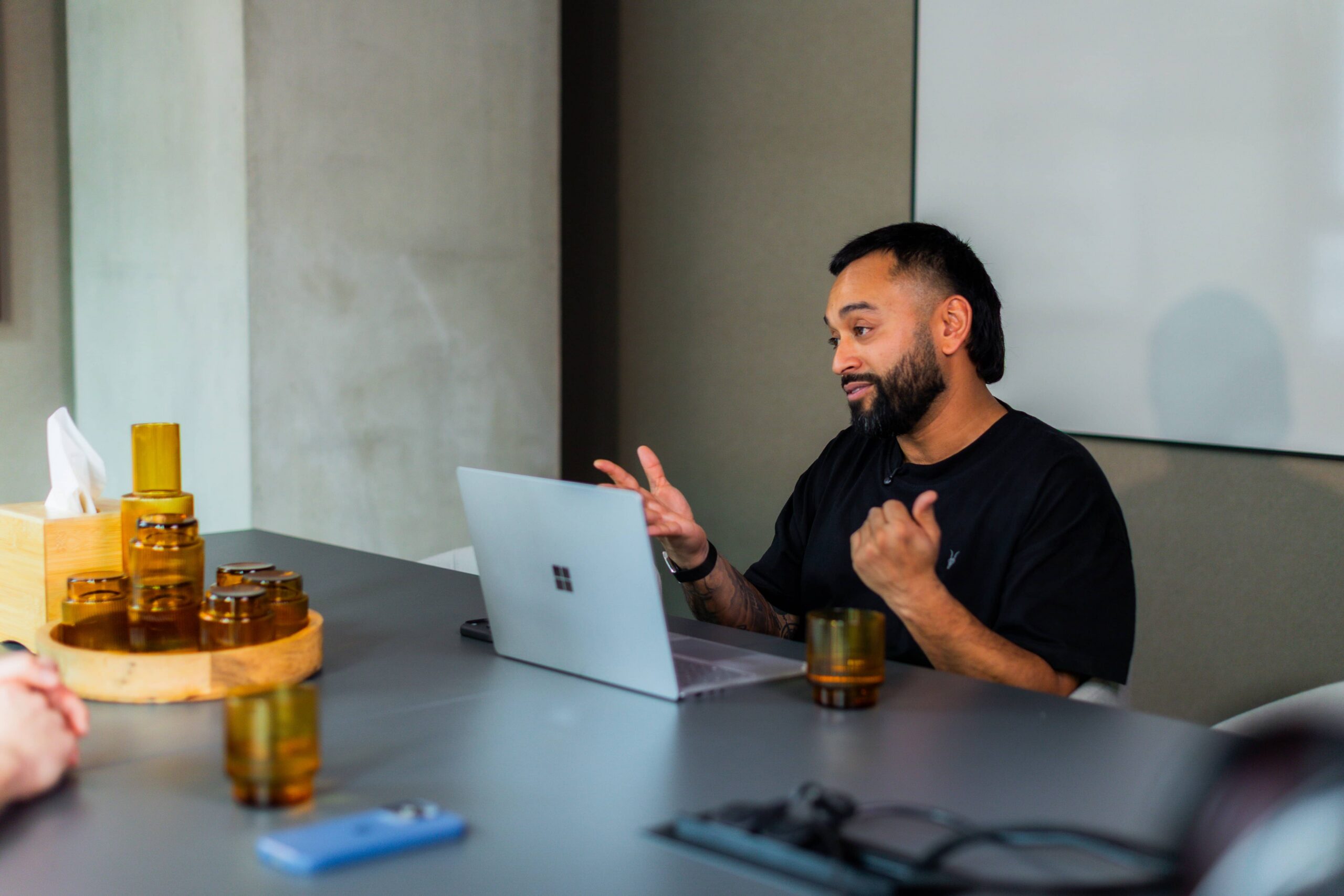 Saeed Bor at a table with a laptop, gesturing as she speaks. Glasses, tissue box, and phone are also on the table.