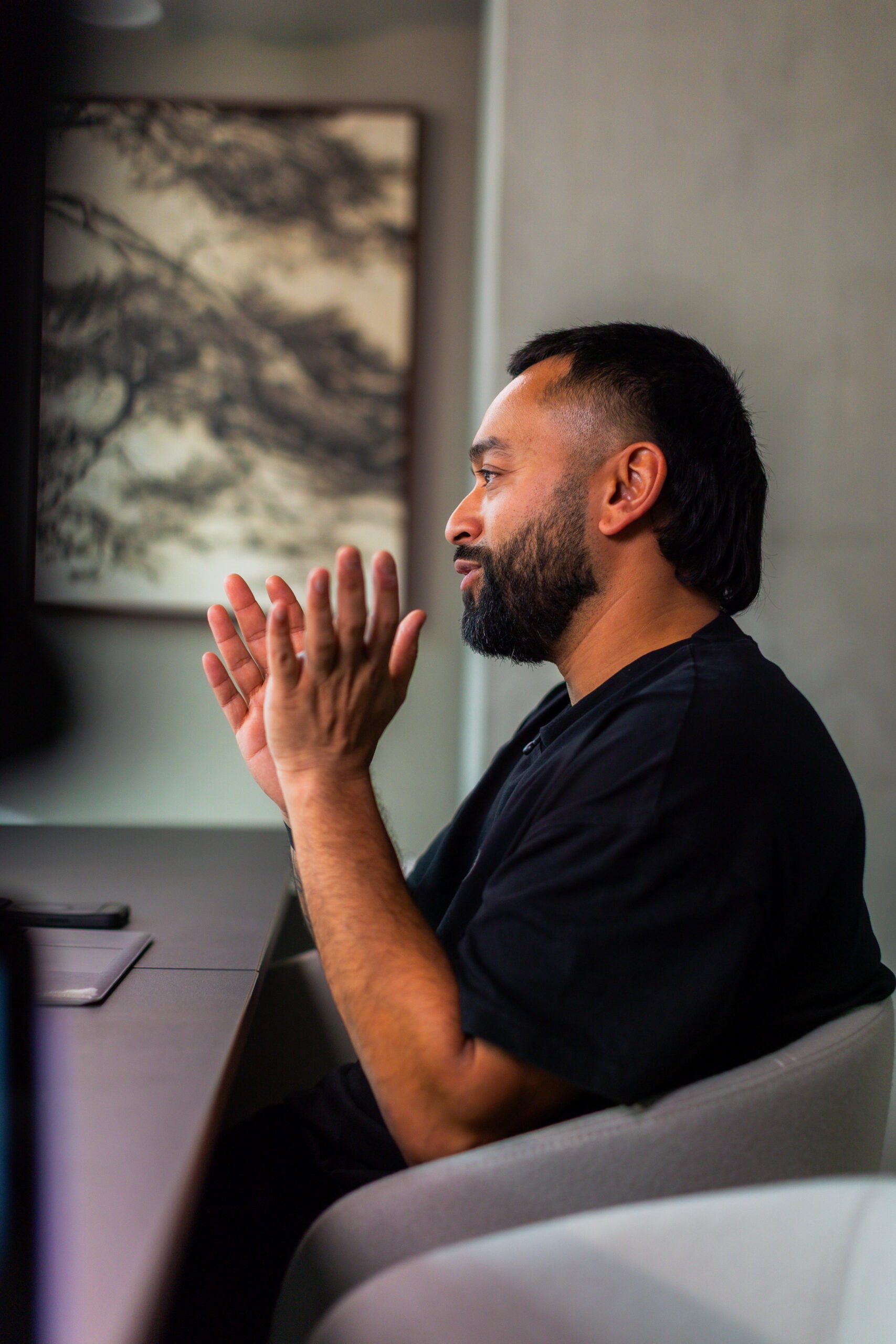 Saeed Bor, bearded, claps at a table with framed artwork on the gray wall in the background.