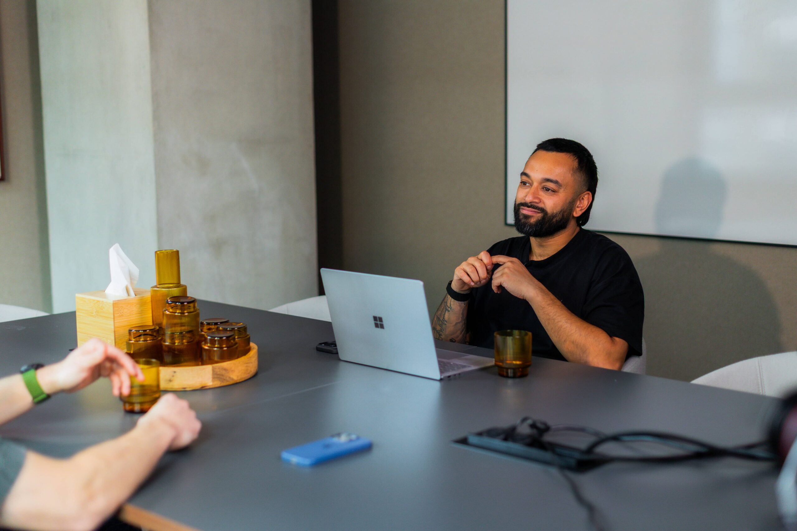 Saeed Bor at a conference table with laptop, glassware, and tissue box, talking to someone off-camera.