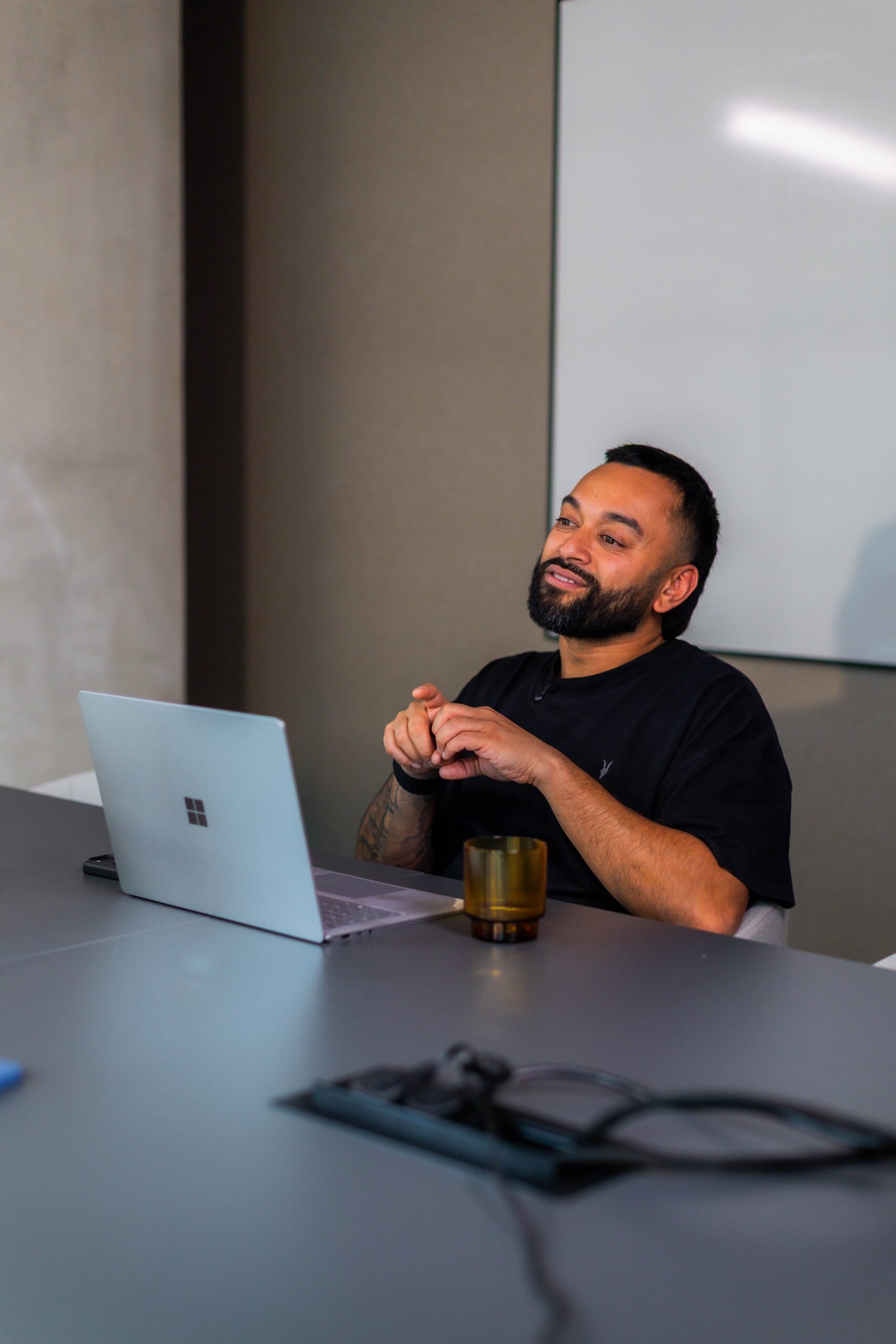 Saeed Bor smiles at a conference table with a laptop and glass, looking upward, whiteboard in background.
