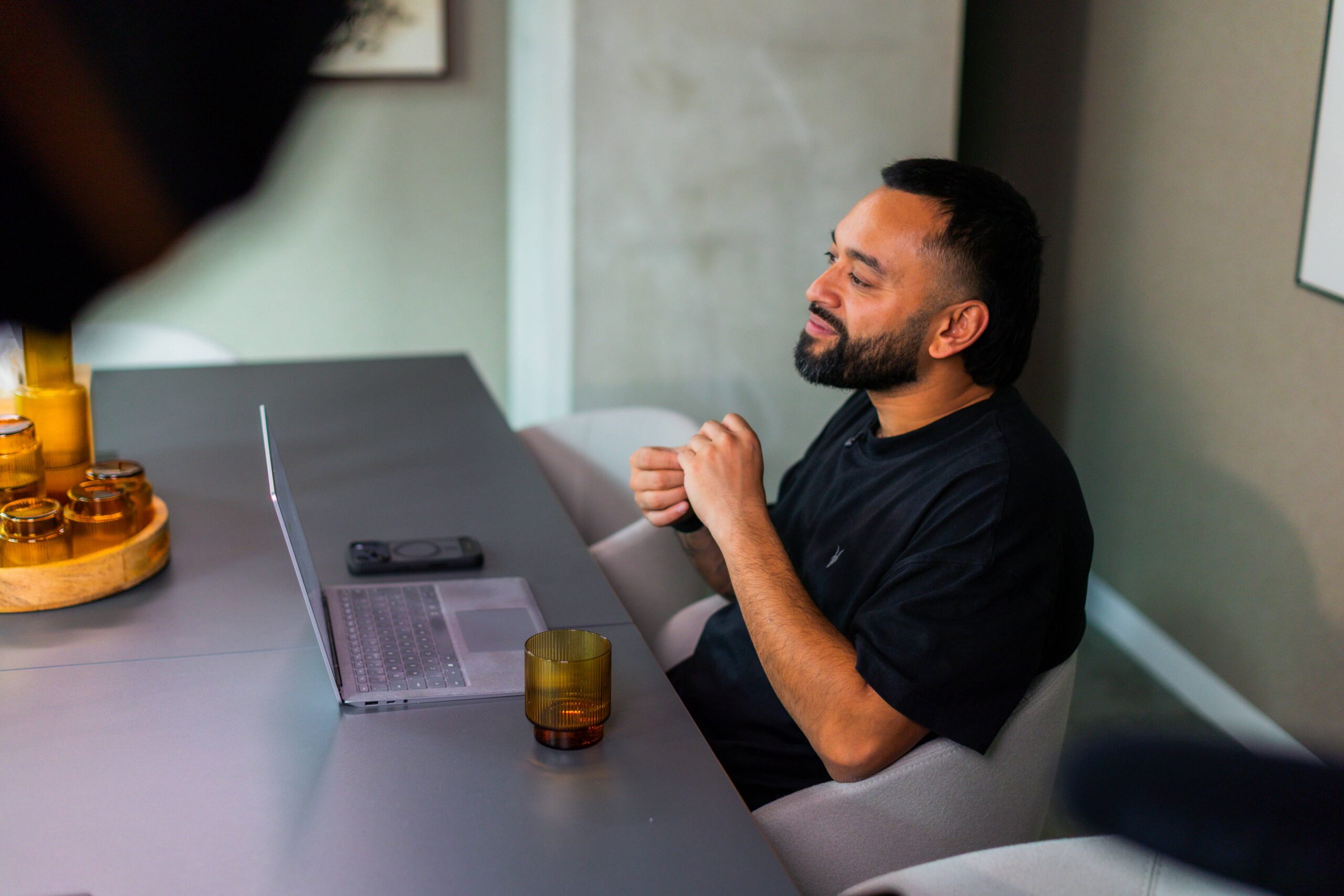 Saeed Bor sits at a table with a laptop, smartphone, and drink, smiling and looking to the side in a modern indoor setting.