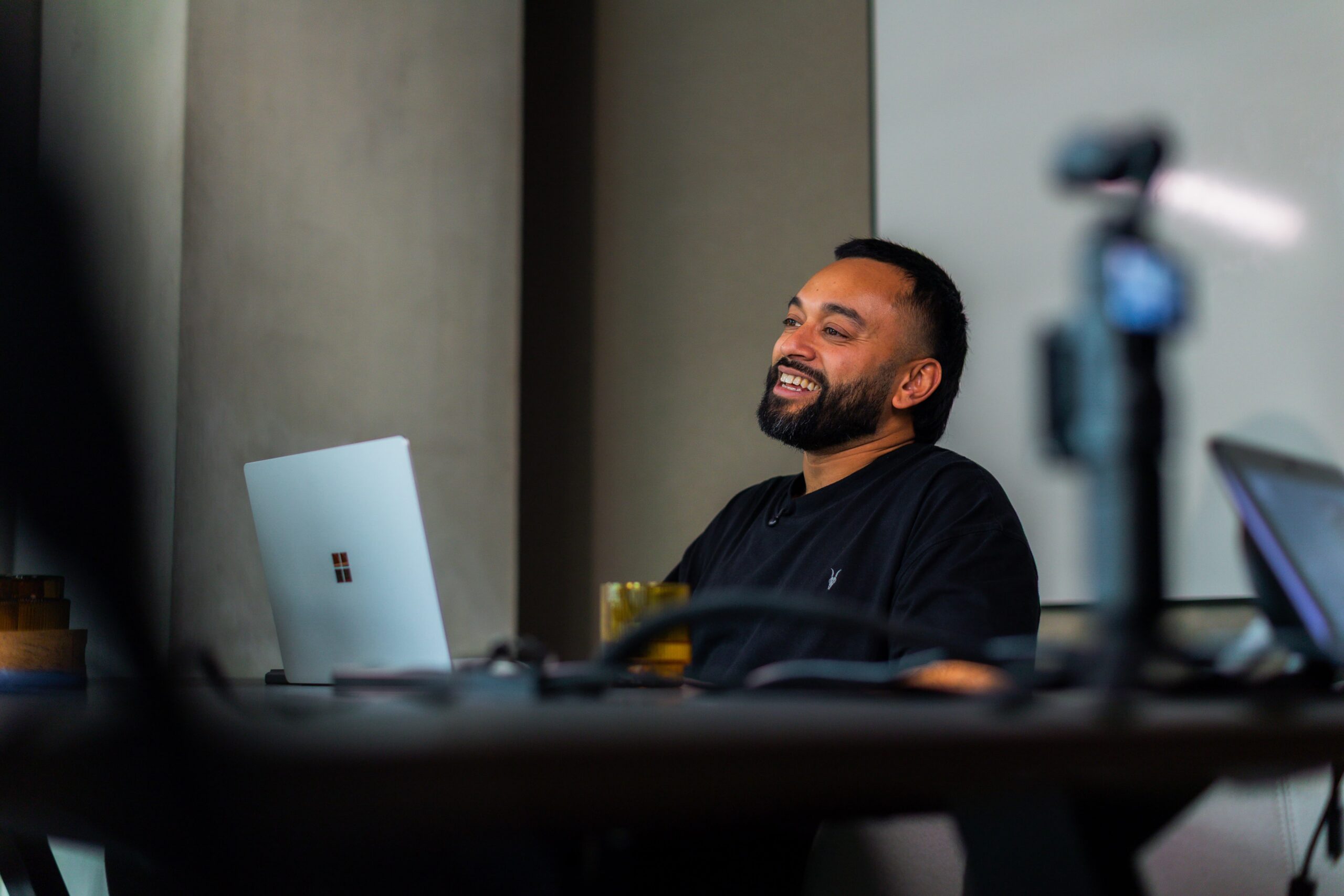 Saeed Bor, a bearded man, smiles while using a laptop at a table; camera and another laptop in the foreground.